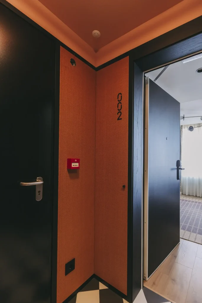 Orange wall corner with a black door ajar, revealing a room with sunlight coming through curtains.