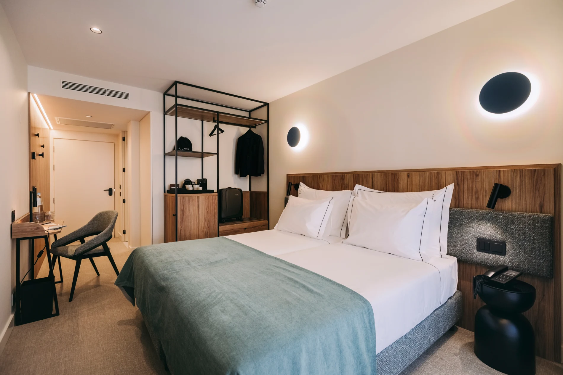 Modern hotel room with a double bed, wall lights, desk, chair, and open wardrobe.