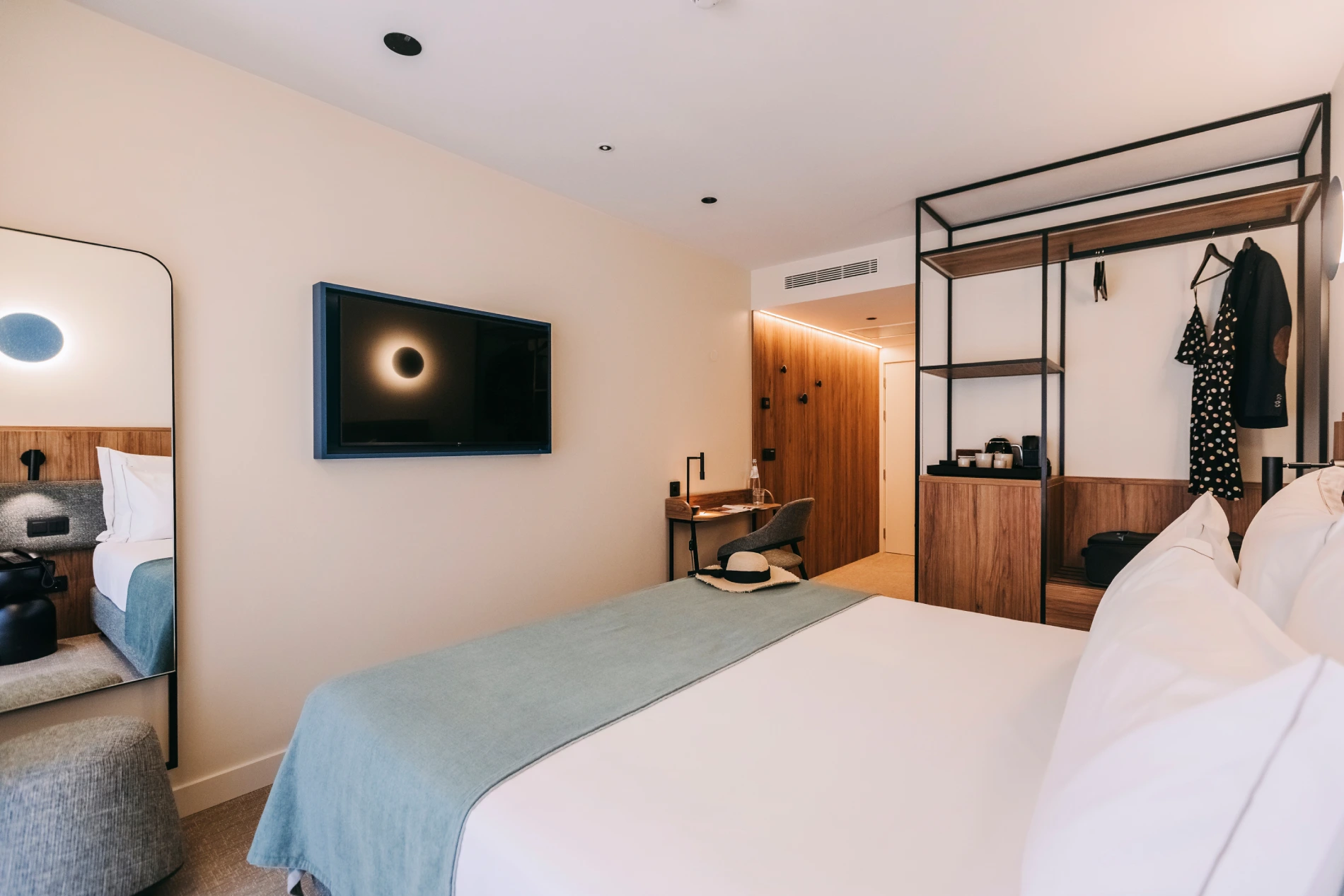 Modern hotel room with a bed, TV, mirror, desk, and open closet with clothes.