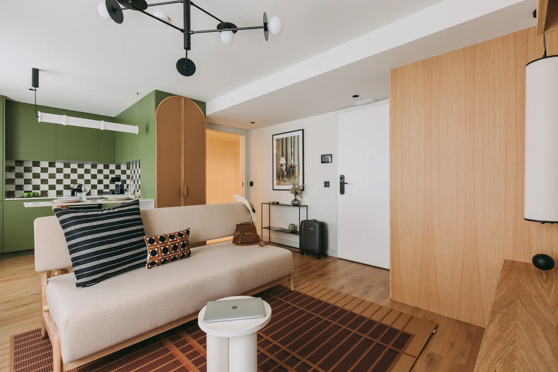 Modern living room with beige sofa, green kitchen, and checkered backsplash.
