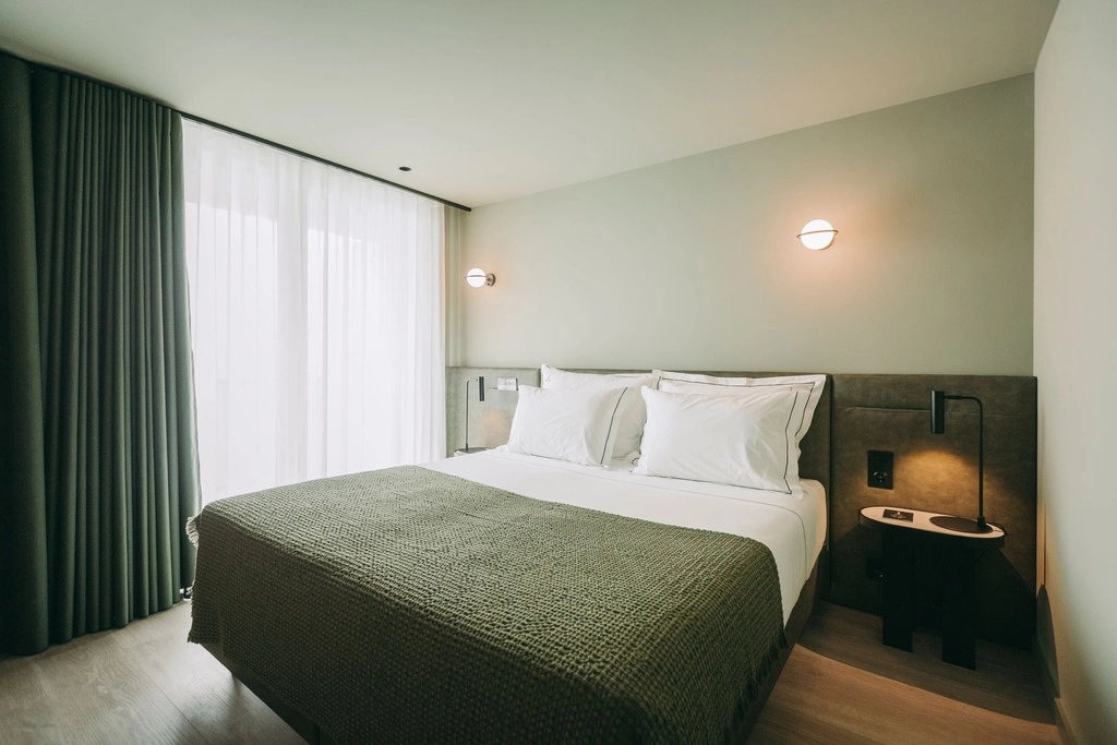 Minimalist hotel room with a double bed, two pillows, modern lighting, and curtains.