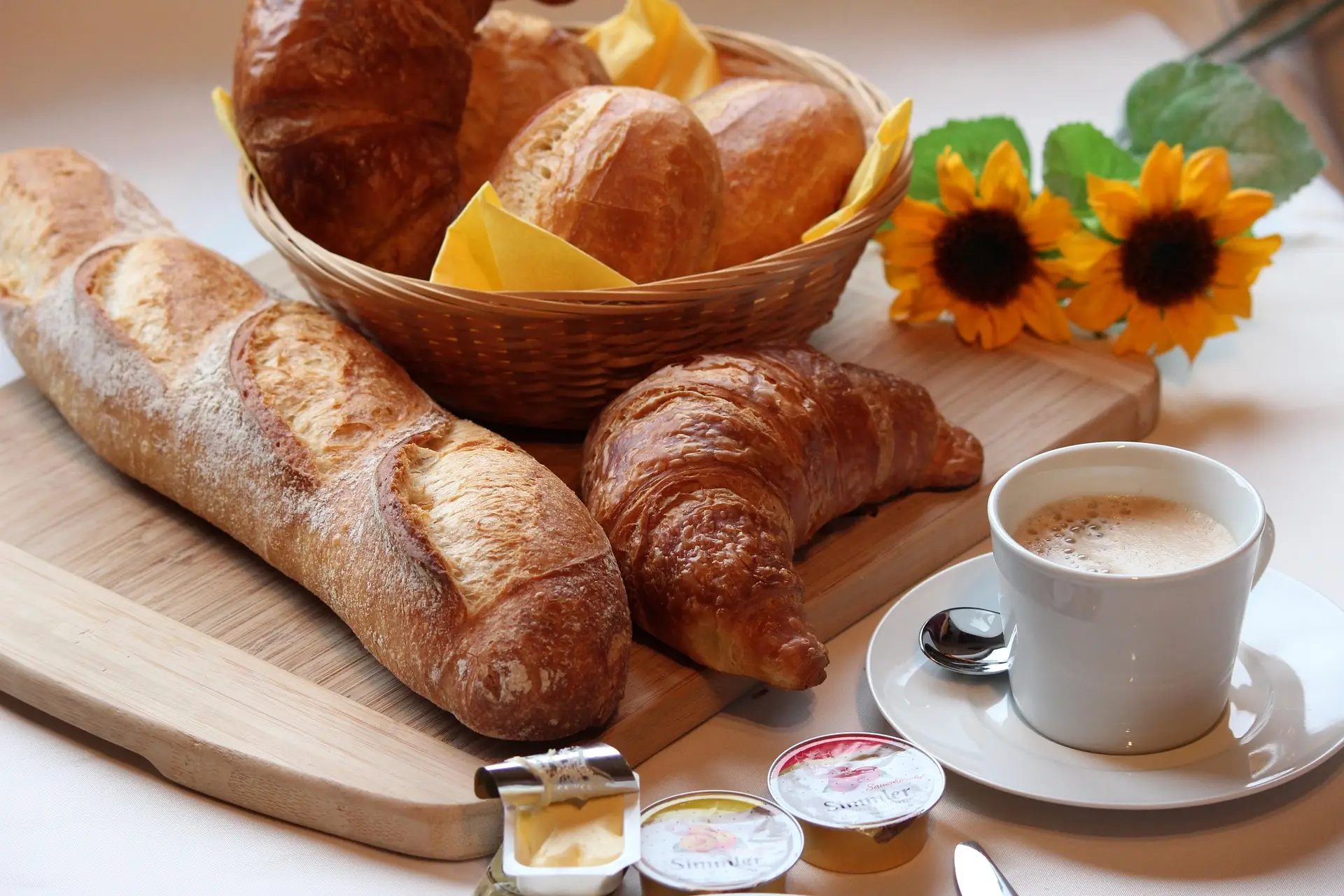 Bread, croissant, coffee, and condiments on a wooden board with sunflowers.