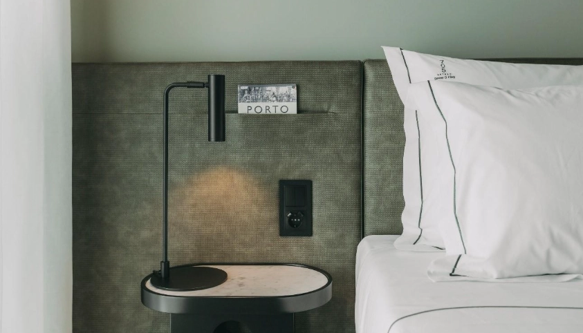 Bedside table with lamp, power outlet, and a card labeled "Porto" next to white pillows.