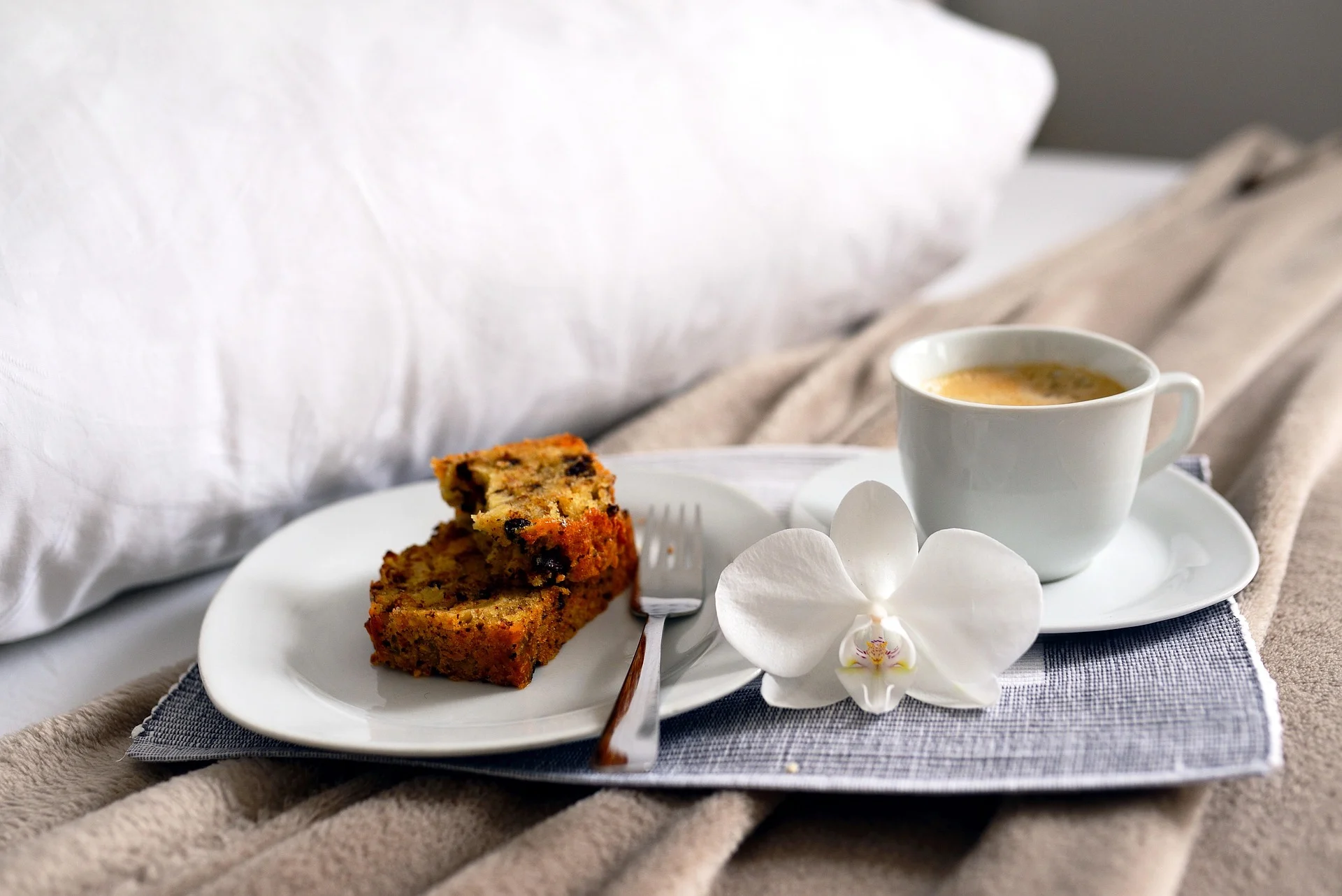 Coffee, cake slices, and an orchid on a bed with beige blanket and white pillow.