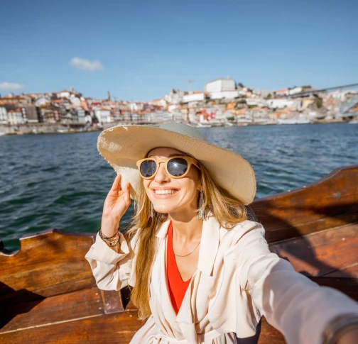 Smiling person in a hat and sunglasses on a boat with a coastal city in the background.
