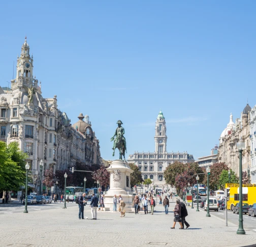 City square with a statue, historic buildings, and people walking on a sunny day.