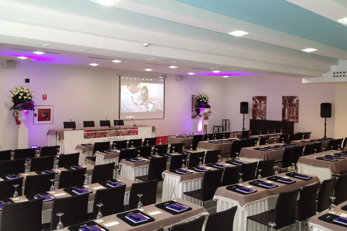 Sala de conferencias con sillas, mesas preparadas, proyector encendido y decoraciones florales.
