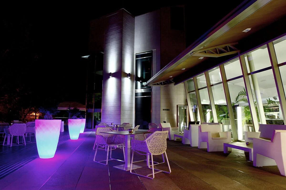 Terraza nocturna iluminada con luces violetas, muebles modernos y plantas cerca de edificio acristalado.