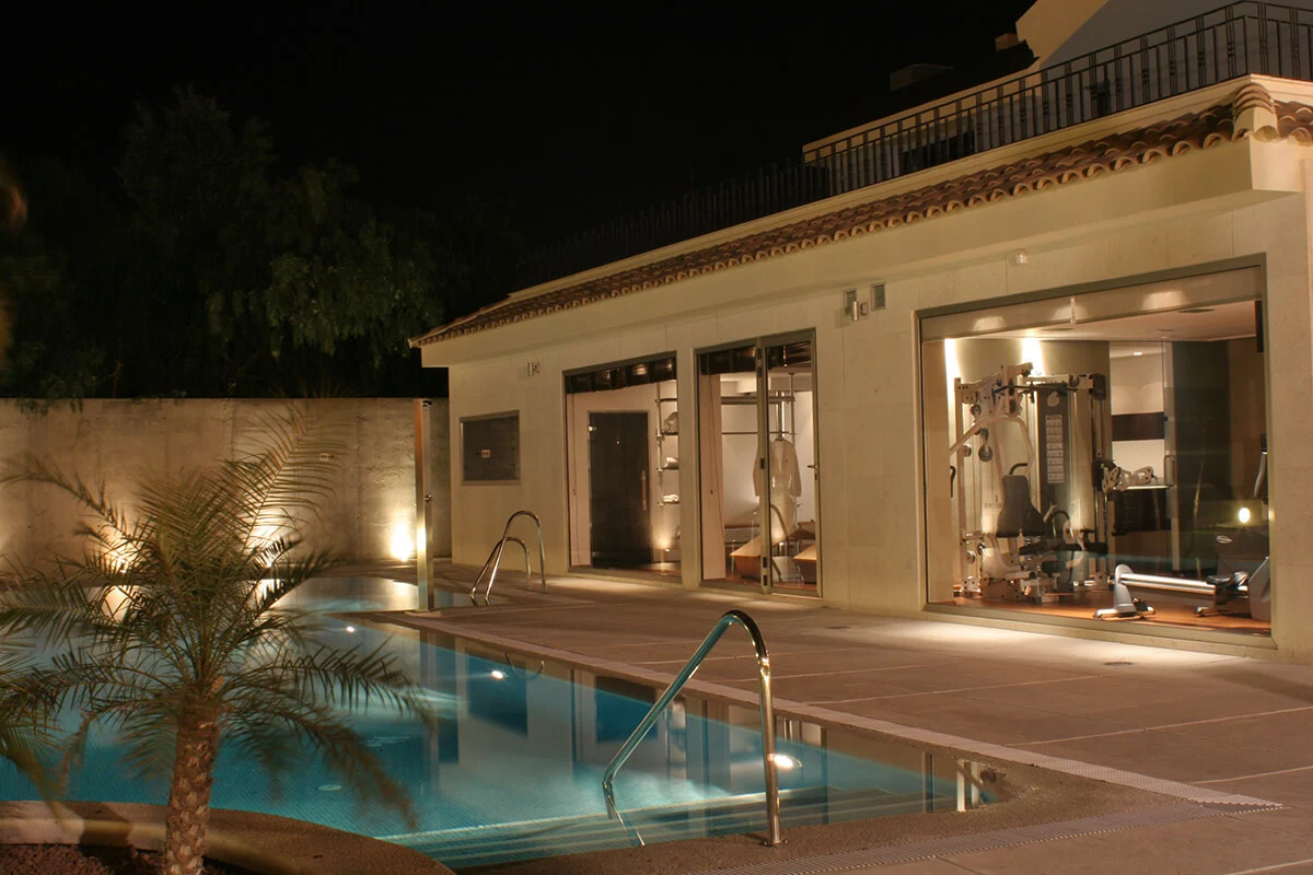Piscina iluminada de noche frente a un gimnasio moderno con grandes ventanales y equipo de ejercicios visible.