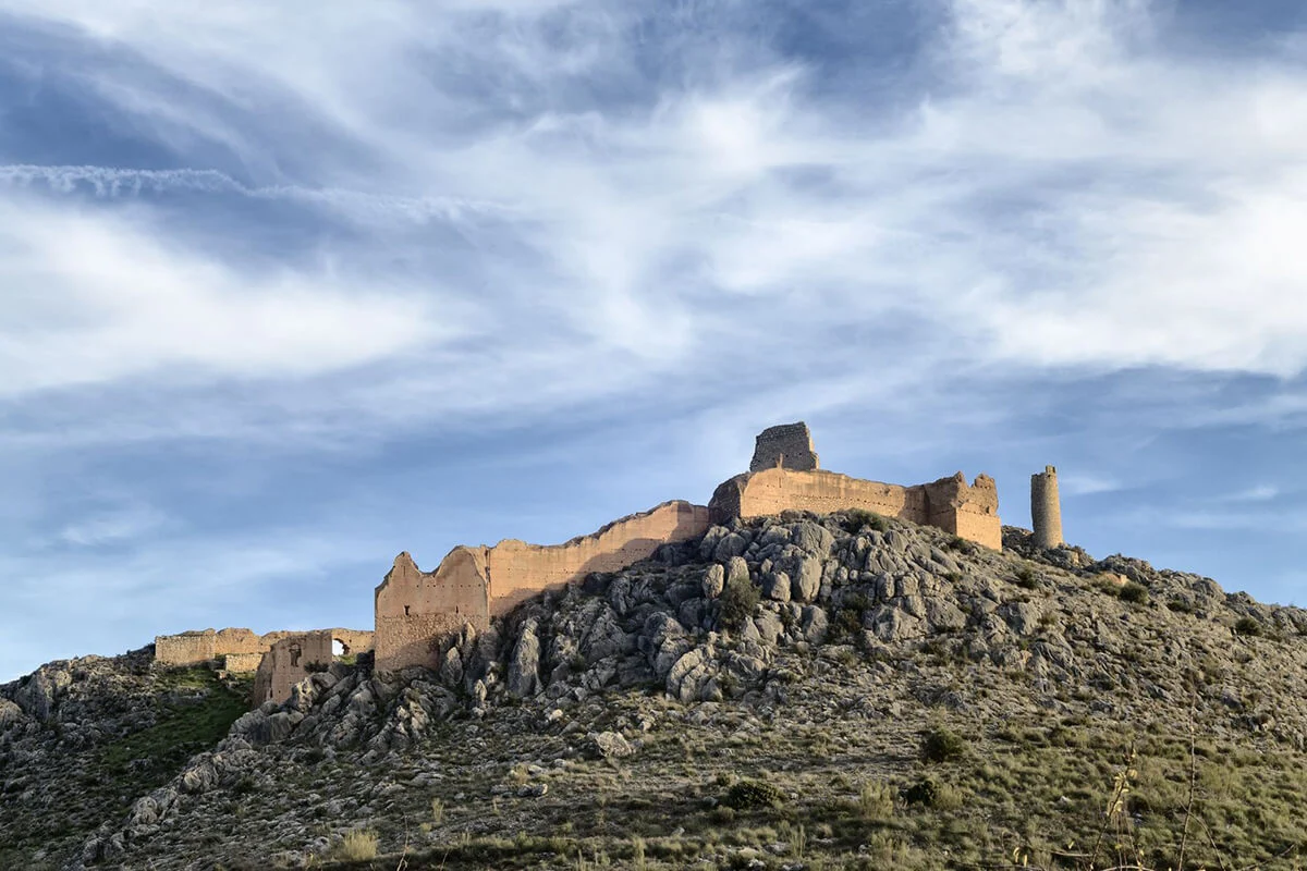 Ruinas de un castillo sobre una colina rocosa, con un cielo azul y nubes dispersas al fondo.