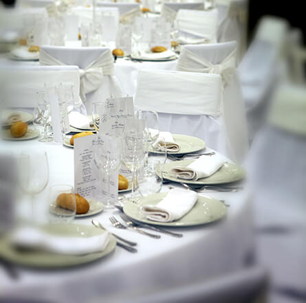 Mesa elegante con platos, copas y servilletas blancas, decorada para un evento especial, con sillas cubiertas.