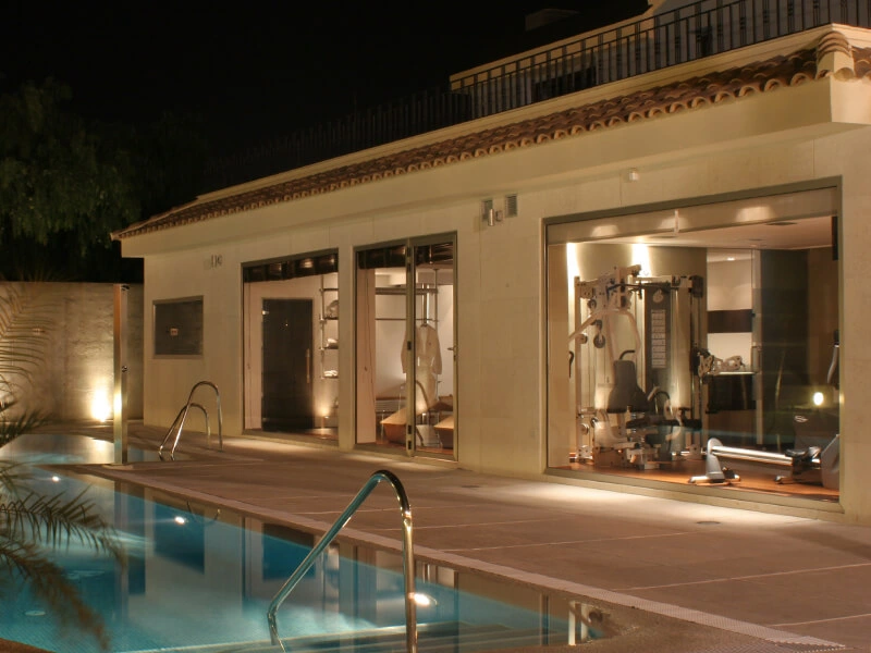 Casa moderna con gimnasio iluminado, vista de piscina en primer plano durante la noche.