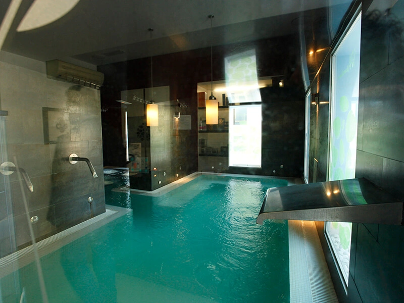 Piscina interior con paredes oscuras, luces colgantes y una cascada de agua en un ambiente moderno y relajante.