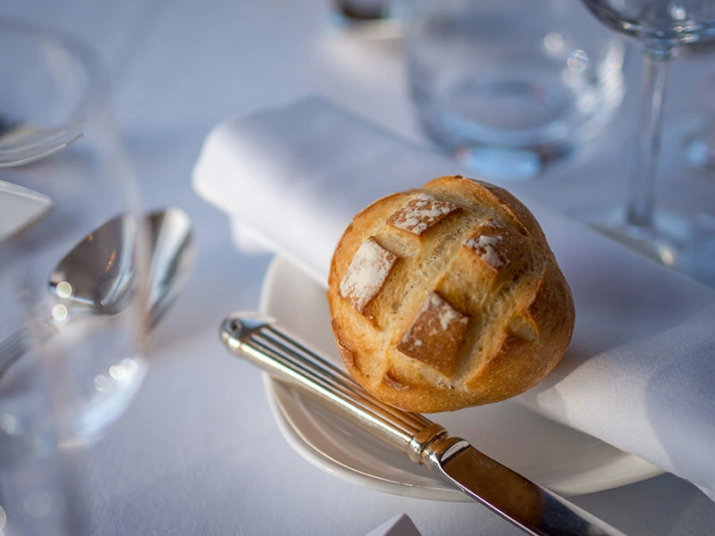 Un pan crujiente en un plato con servilleta, cuchillo de plata, copa de vidrio y mantel blanco elegante de fondo.