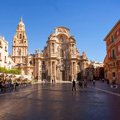 Catedral barroca con plaza amplia, personas caminando y edificios históricos alrededor bajo un cielo despejado.