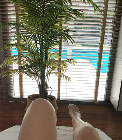 Piernas relajadas en interior junto a ventana con persianas, planta y vista a piscina. Luz natural ilumina la escena.