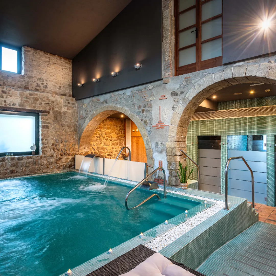 Piscina interior de spa con chorros de agua y cascada, rodeada de paredes de piedra y arcos, en un ambiente cálido y relajante.