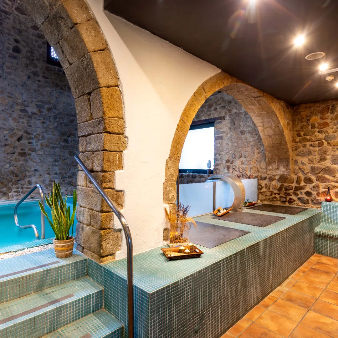 Zona interior de spa con paredes de piedra y arcos, escaleras revestidas de mosaico y área de relajación junto a una piscina con cascada de agua.