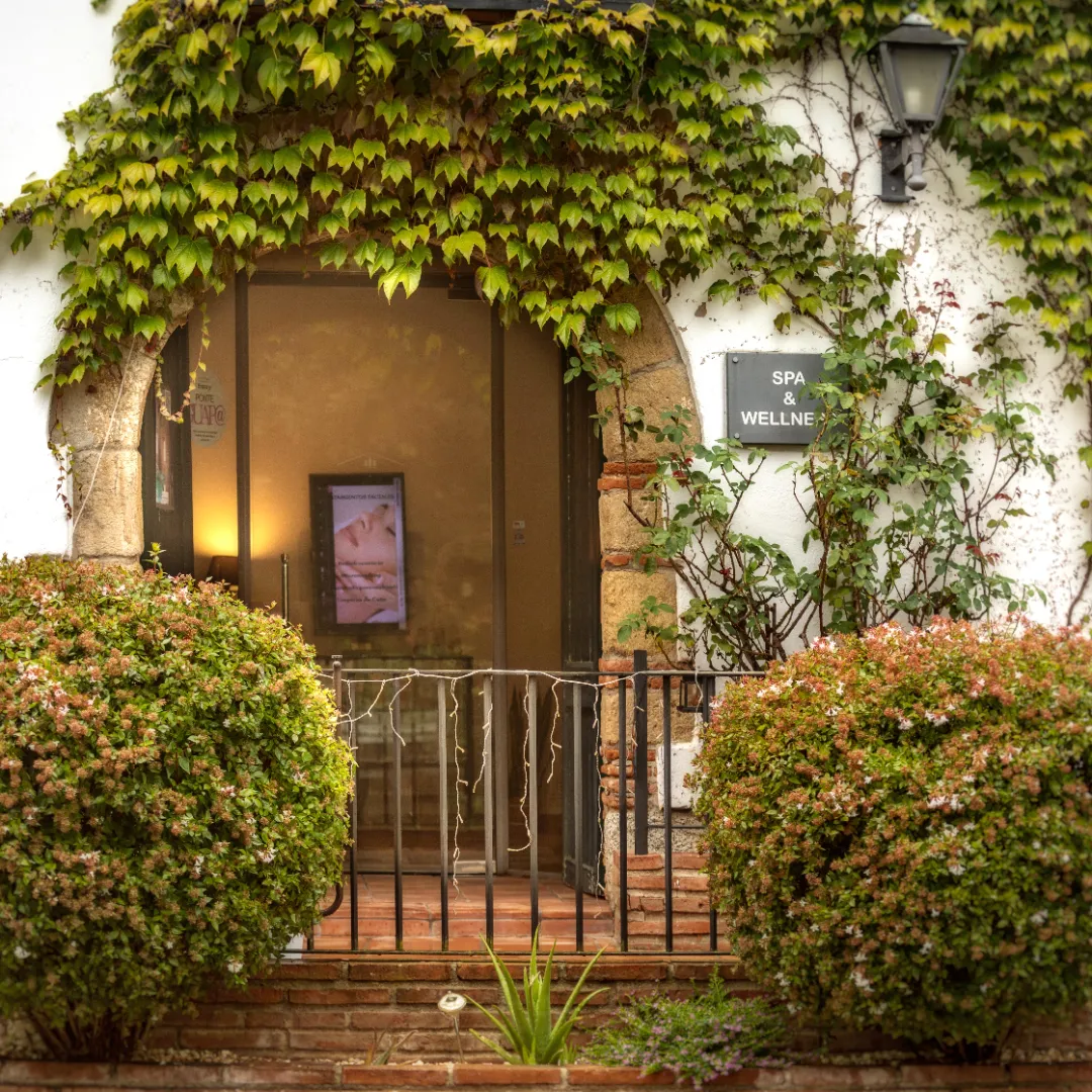 Entrada de un spa y centro de bienestar con una puerta de vidrio rodeada de plantas trepadoras y arbustos, en una fachada rústica de piedra y ladrillo.