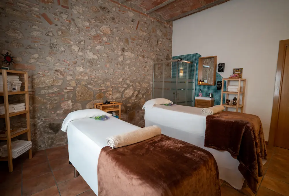 Sala de masajes con dos camillas, toallas y decoración en un ambiente de pared de piedra y estanterías con productos.