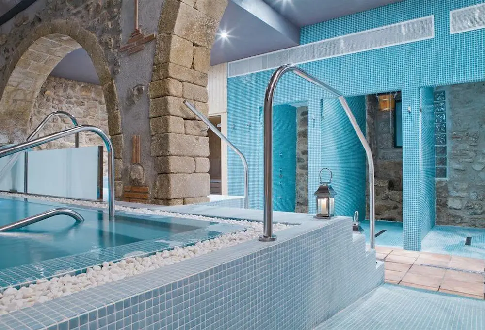 Piscina interior con mosaicos azules, arcos de piedra y barandillas de acero. Linterna decorativa en un rincón.
