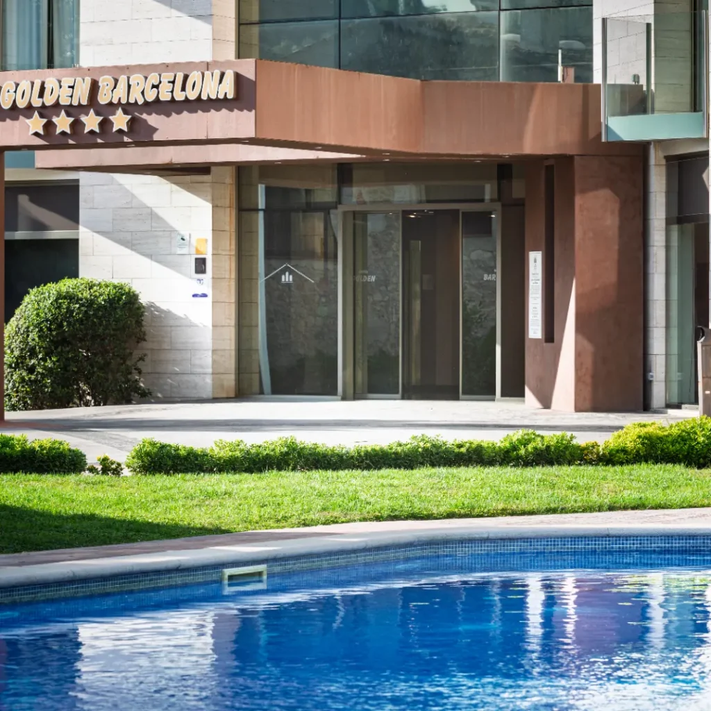 Una piscina frente a un edificio moderno, rodeada de áreas verdes y tumbonas.