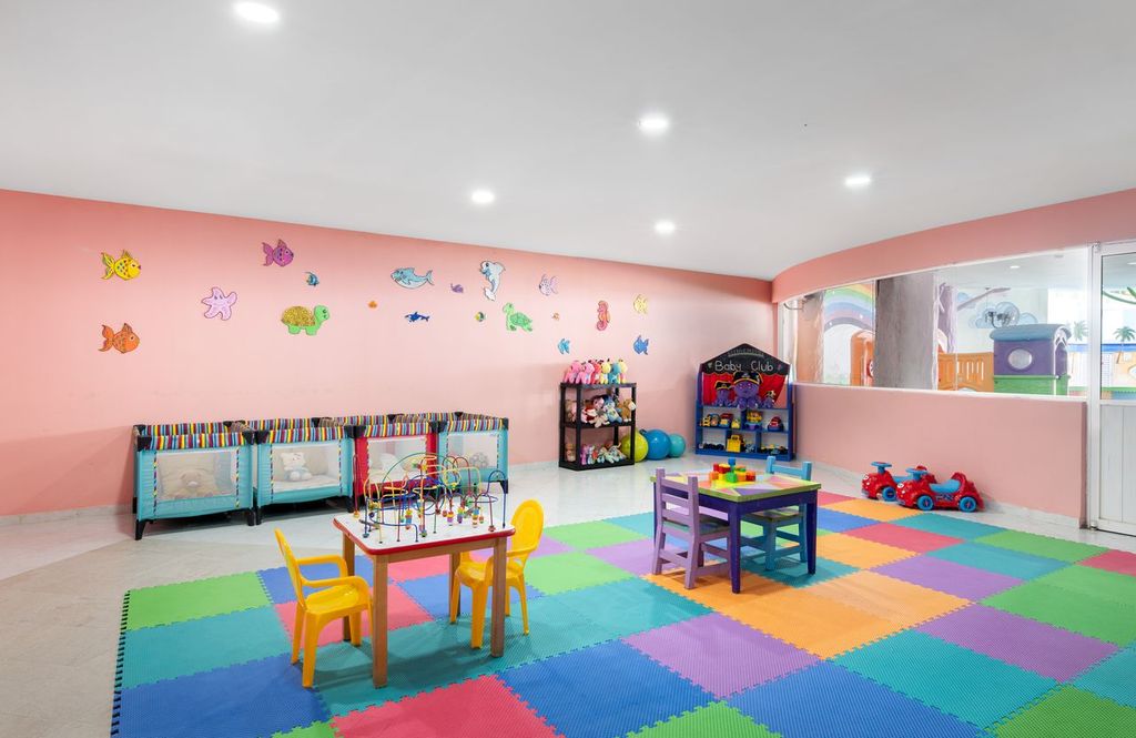 Colorful children's playroom with tables, chairs, toys, and wall decorations on soft floor mats.