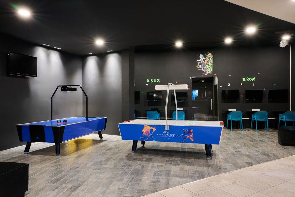 A modern game room with air hockey tables, Xbox stations, and blue chairs, lit by ceiling lights.
