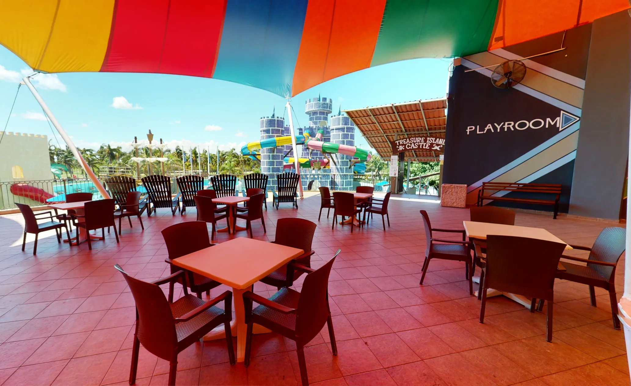 Outdoor seating with colorful shade, tables and chairs, view of water slides and building labeled "Playroom."