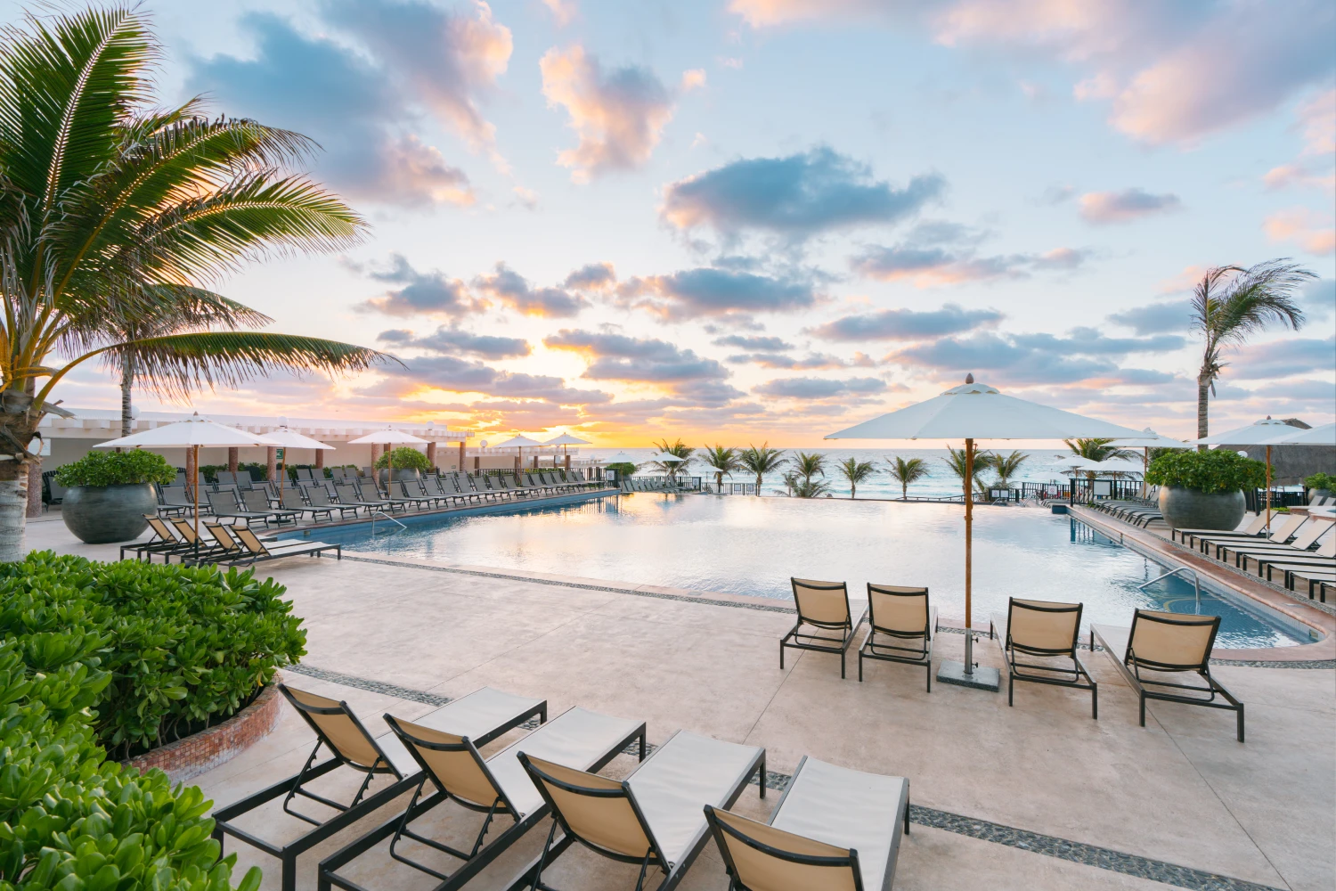 Poolside loungers and umbrellas overlook the ocean at sunset, with palm trees and a partly cloudy sky.