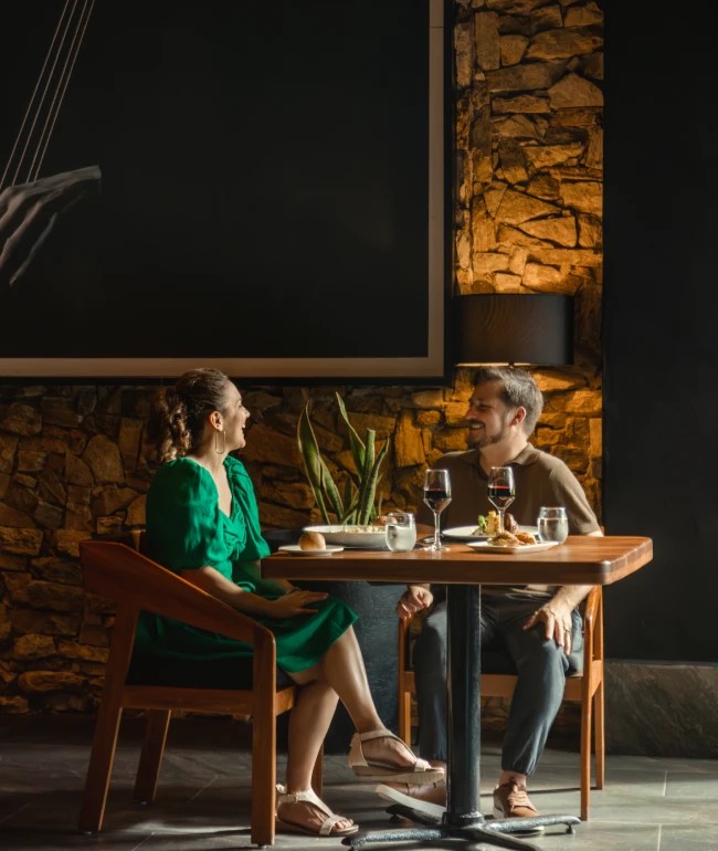A couple enjoys wine at a cozy restaurant table, surrounded by stone walls and a plant.