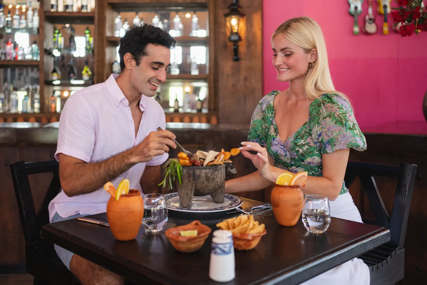 A couple enjoys a meal with drinks at a restaurant, featuring a vibrant pink wall and a variety of bottles in the background.