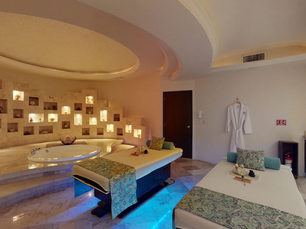 Luxurious spa room with two massage beds, a round jacuzzi, decorative shelves, and a robe on the wall.