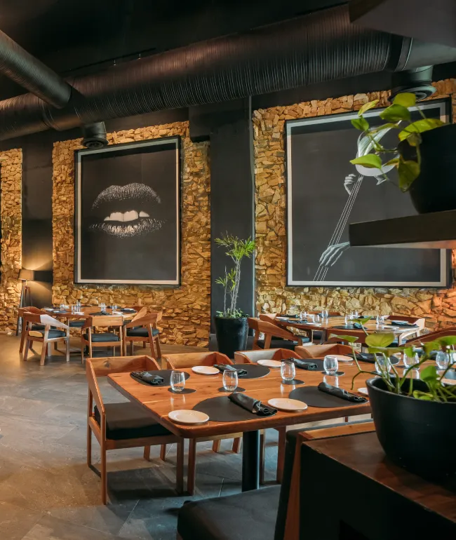 Cozy restaurant with wooden tables, stone walls, artistic wall art, and lush green plants.