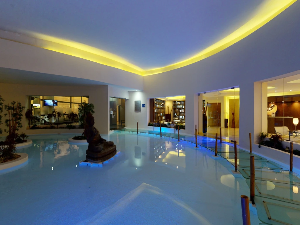 Modern interior with a curved pool, glass bridges, and soft lighting. Statues and plants accent the serene space.