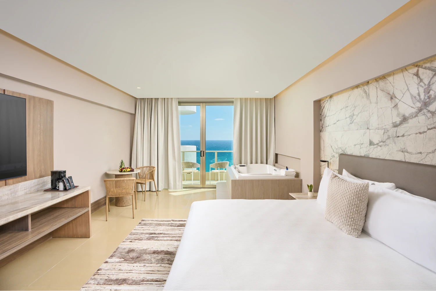 Hotel room with a bed, TV, table, and chairs. Balcony with ocean view, jacuzzi near window, neutral tones.