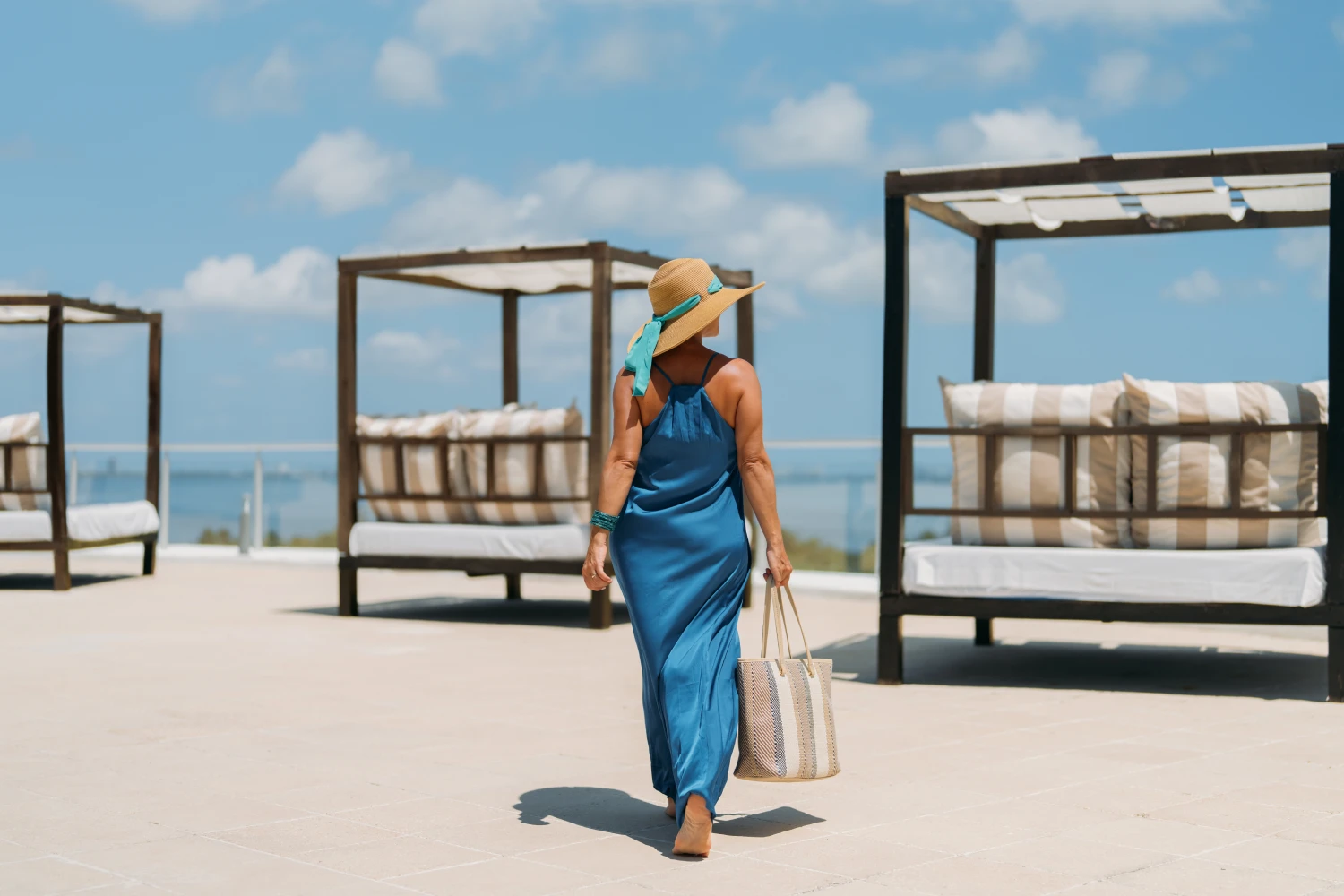 Woman in a blue dress and hat walking by outdoor cabanas under a blue sky.