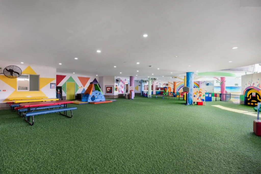 Colorful indoor playground with green turf, benches, and climbing area. Geometric wall art and play structures visible.