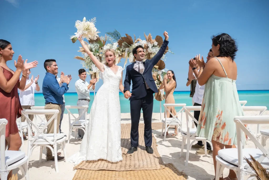 Bride and groom celebrate with guests on a sunny beach, surrounded by chairs and floral decorations.