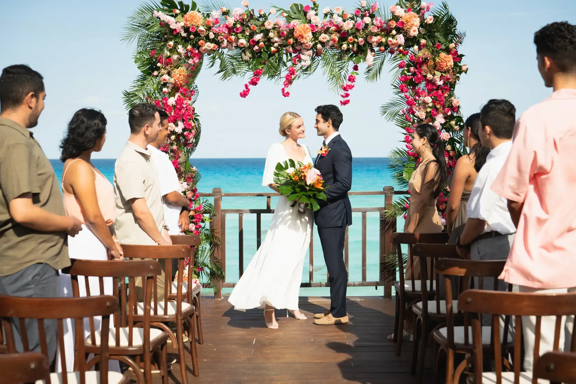 Couple getting married on a beach deck, under a floral arch, surrounded by guests, with the ocean in the background.