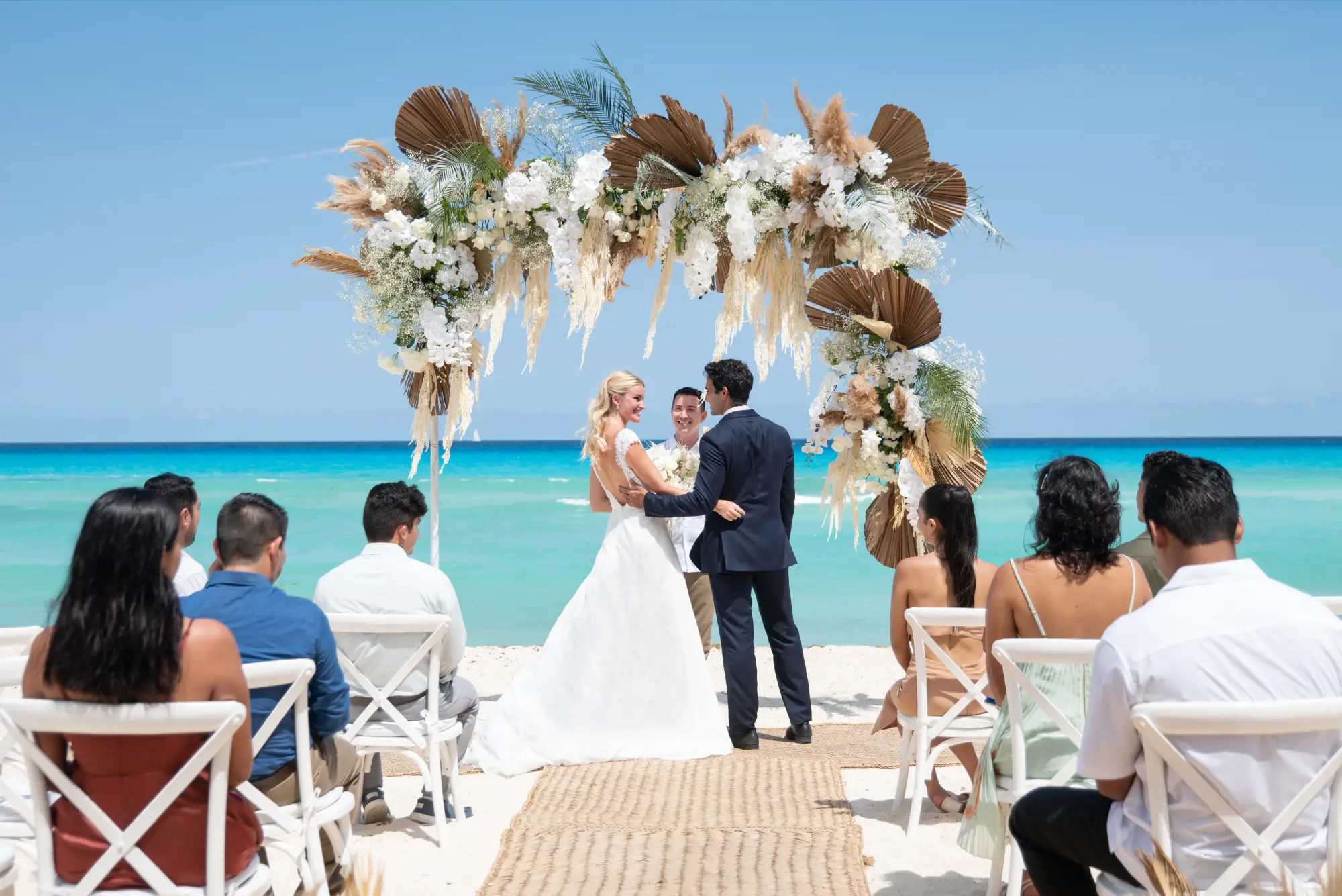 Beach wedding ceremony with a floral arch. Couple stands before guests seated on the sand, ocean in the background.