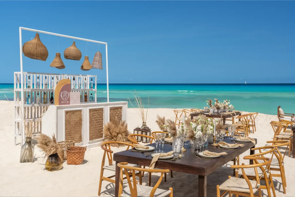 Beachside dining setup with wooden furniture, decorative plants, and a bar under a clear blue sky.