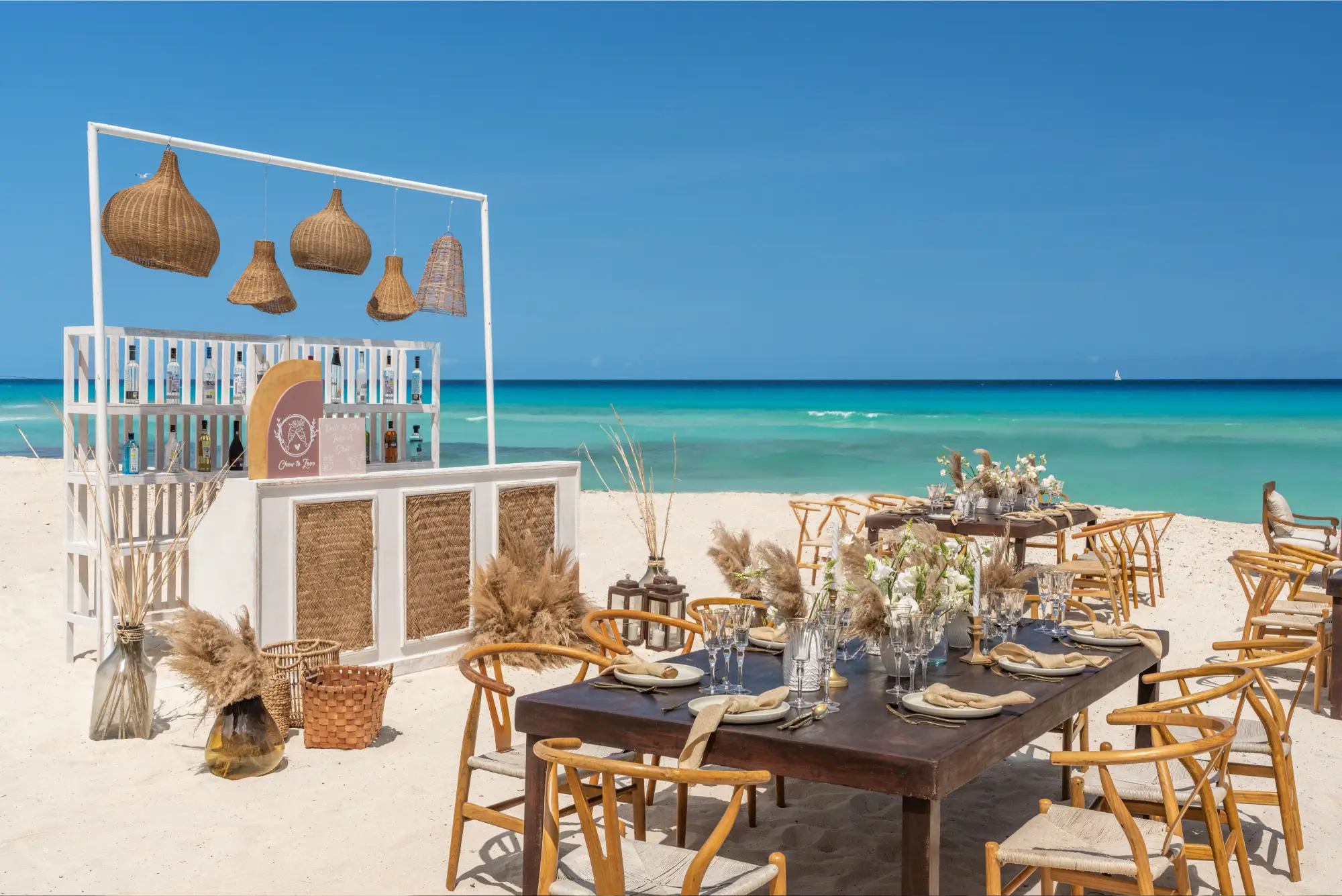 Beachside dining setup with wooden furniture, decorative plants, and a bar under a clear blue sky.