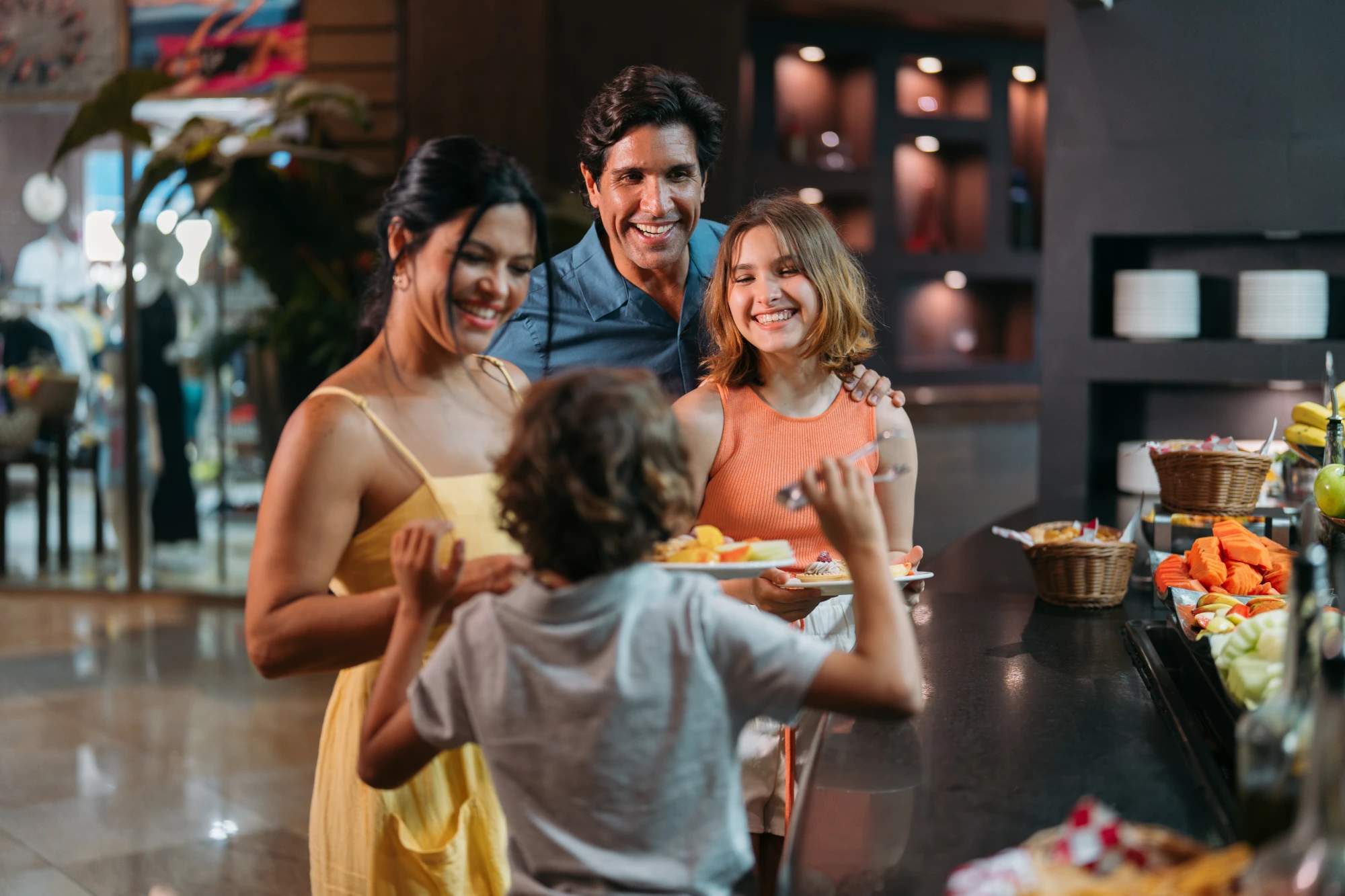 Family smiling and selecting food from a buffet table, holding plates with fruit and pastries.