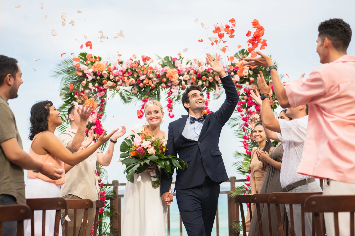 Bride and groom walk under a floral arch, smiling as guests throw petals and cheer.