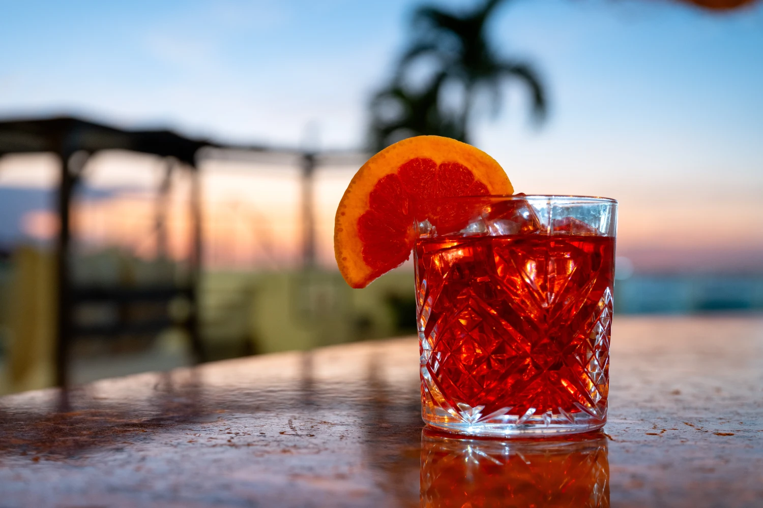 A cocktail with an orange slice on a textured table, sunset and palm tree blurred in the background.