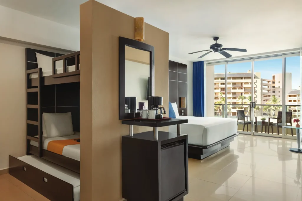 Hotel room with bunk beds, queen bed, coffee maker, and a balcony view of a building with palm trees.