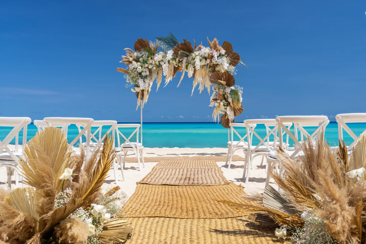 Beach wedding setup with wooden chairs, a floral arch, and an aisle leading to the turquoise sea under a clear blue sky.
