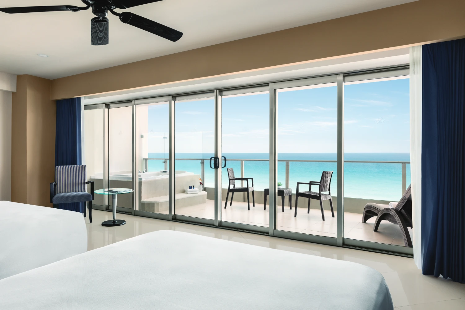 Hotel room with ocean view, glass doors, balcony with chairs, and a bathtub. Bright and airy space.