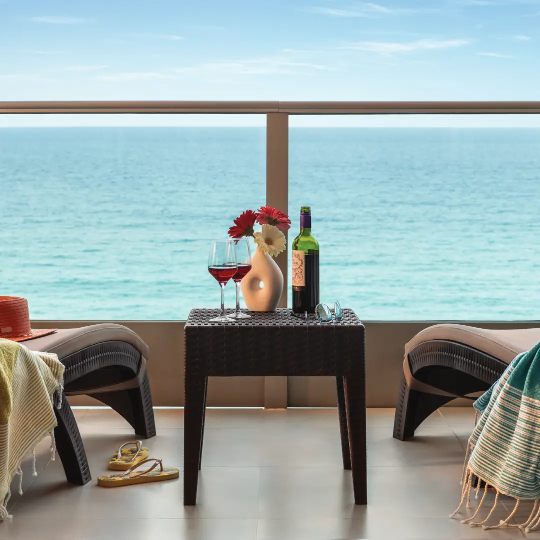 Balcony with ocean view, table with wine, glasses, flowers, sunglasses, sandals, two loungers with towels.